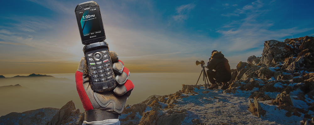 Background shows a photographer taking a photo over a mountain with a hand in the foreground holding up a kyocera phone