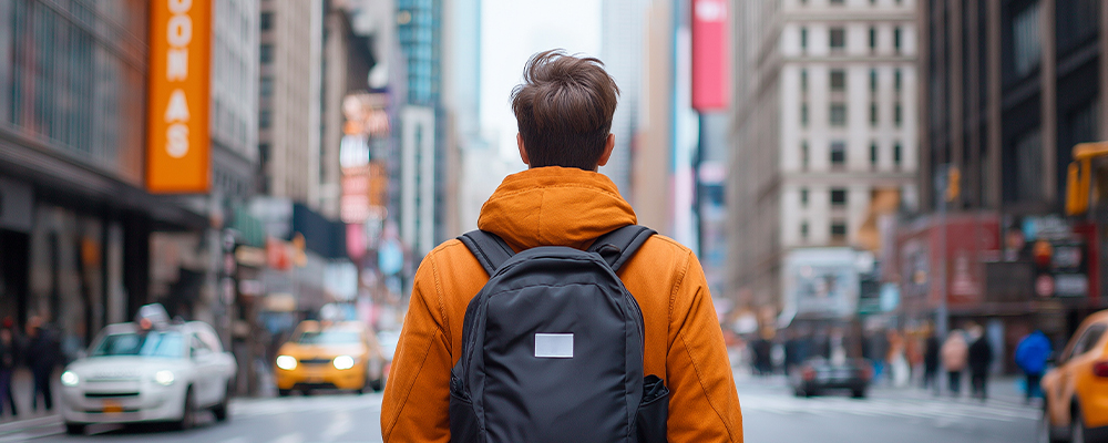 A man wearing an orange hoodie and a black backpack is seen from behind as he navigates through a busy city street.